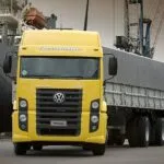 Ein gelber Lastwagen der Marke Volkswagen Constellation steht vor einem Frachtschiff im Hafen. Der LKW hat eine große, silberne Kühlergrill und eine geschlossene Plane auf seinem Anhänger. Im Hintergrund sind Hafenkräne und weitere Frachtschiffe zu sehen.